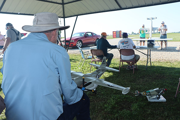 Man with remote control plane under a pavilion at a field event.
