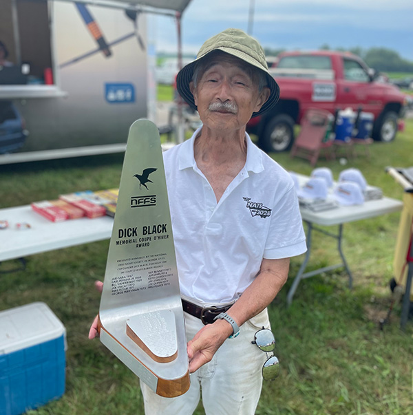 Elderly man holding a gray and wooden trophy outdoors, wearing a sunhat and white shirt.