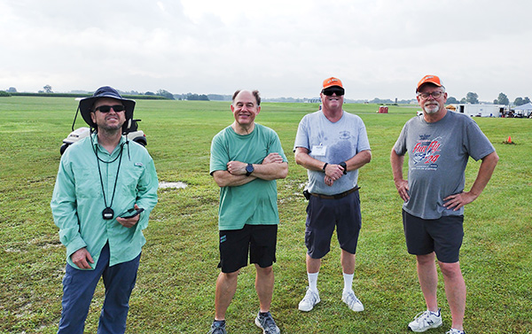 Four men standing on grass, casually dressed, with cloudy sky overhead.