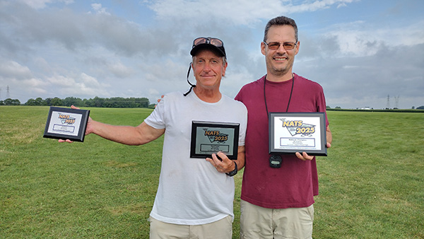 Two men outdoors smiling, each holding awards, standing on a green field under a cloudy sky.