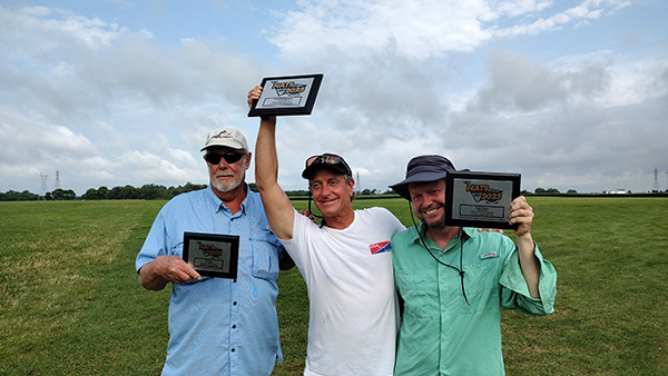 Three smiling men holding framed certificates in a grassy field.