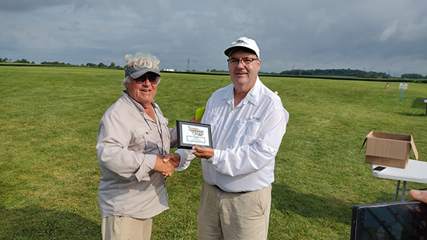 Two men outdoors, one handing a plaque to the other, with a grassy field background.