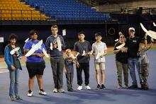 Hope, Joshua, and Caleb Finn (right) of J & H Aerospace pose with some of the youth competitors that they mentor. All are holding gliders from J & H Aerospace. Photo credit: Mai Le Group with model airplanes in a stadium setting.