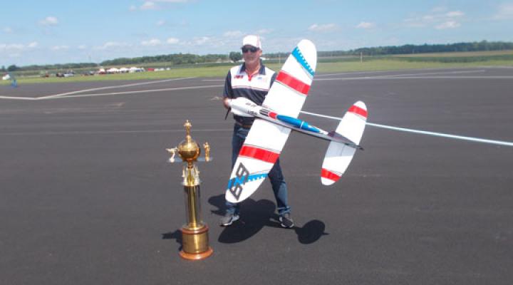 Man with model airplane, standing beside large trophy on airfield.