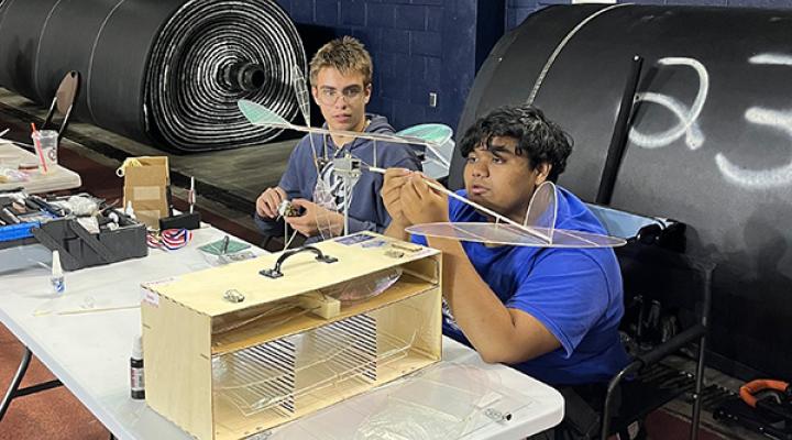 Two people assemble a model aircraft at a table, large rolls in the background.