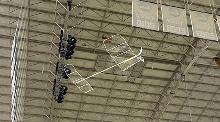 Model airplane flying indoors near a high ceiling with lights and signs.