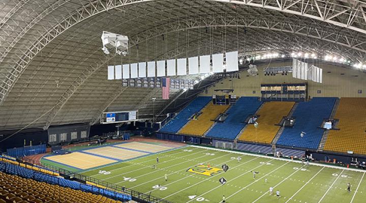 Indoor football stadium with players on the field and rows of blue and yellow seats.