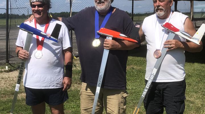 Three men with medals holding model airplanes on a sunny day.