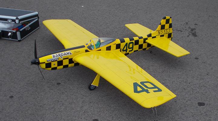 Yellow model plane with checkered black details and a propeller, on a gray surface.