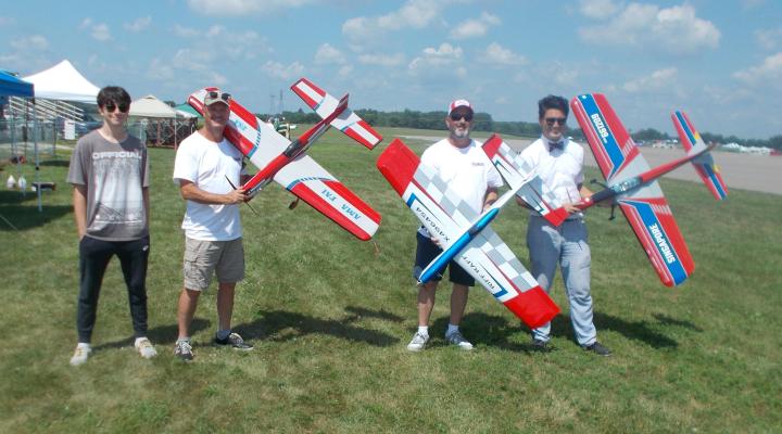 Group of four men outdoors holding red and blue model airplanes.