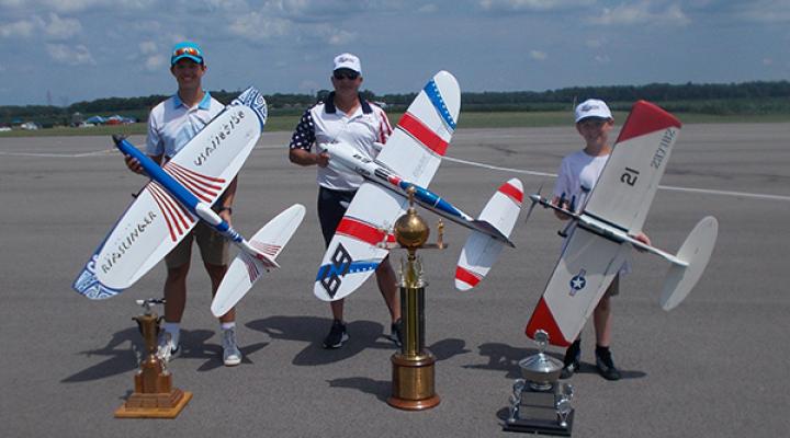 Three people outdoors with large model airplanes and trophies.