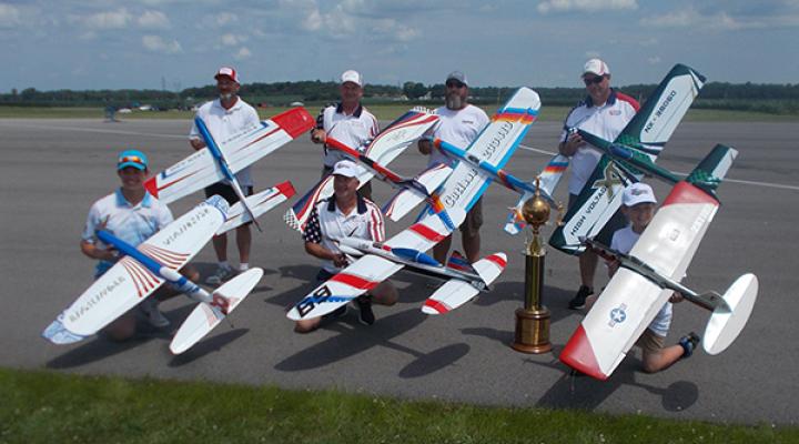 Six people with model airplanes and trophy on an airfield.