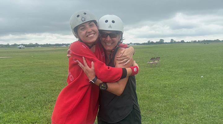 Two people in helmets hugging on a grassy field, smiling under a cloudy sky.