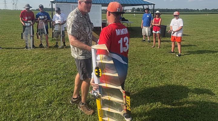 Two men shaking hands at a model airplane event with spectators nearby.