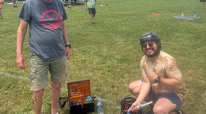 Two people with helmets outdoors, assembling a model airplane on grassy field.