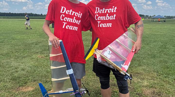 Two people in red shirts holding model planes on grass under a blue sky.