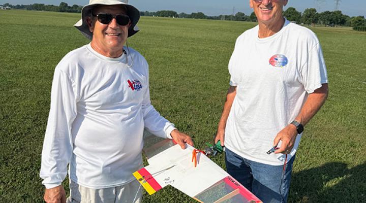 Two men smiling, holding a model glider on a grassy field.