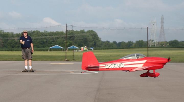 Red model plane flying low near a person on an airfield.