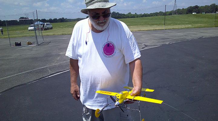 Man holding a yellow model airplane on a sunny day at an airfield.