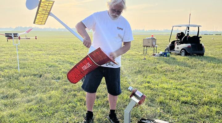 Man assembling red and white model plane on grassy field at sunrise.