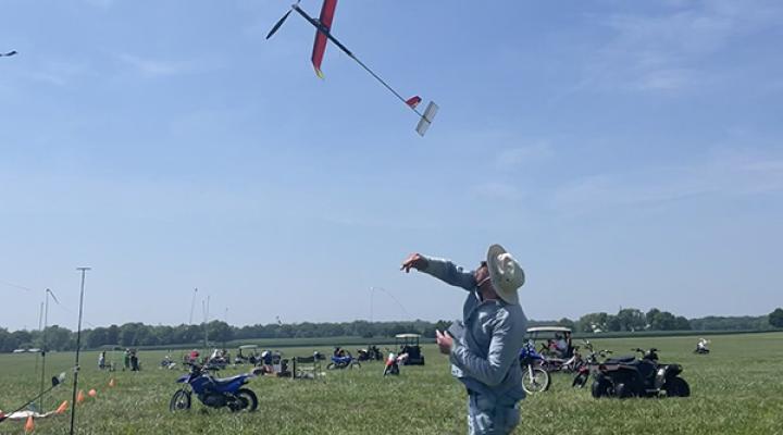 Man launching model glider in grassy field under clear blue sky.