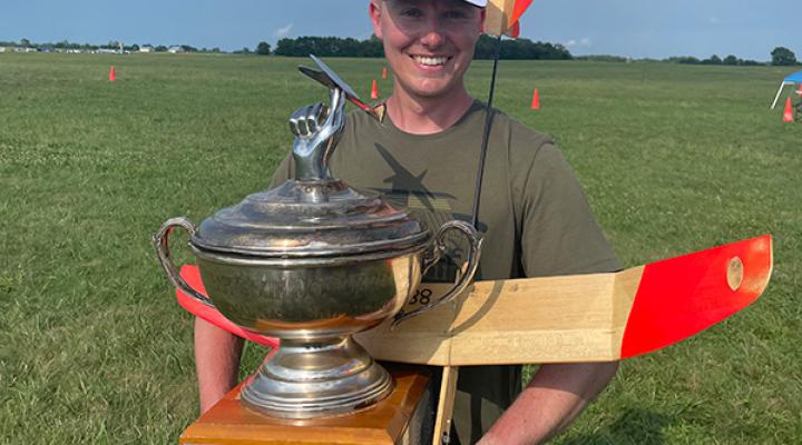 Man holding large trophy and model plane on grassy field.