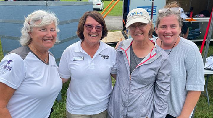 Four women smiling outdoors, wearing casual summer clothing.