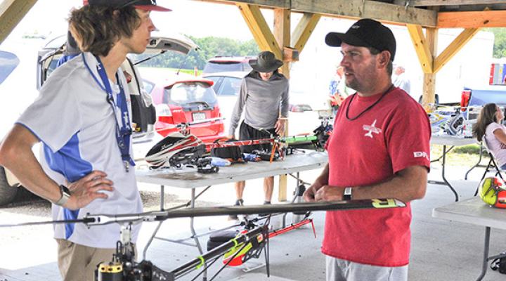 Two men discuss under a pavilion beside RC helicopters on tables.