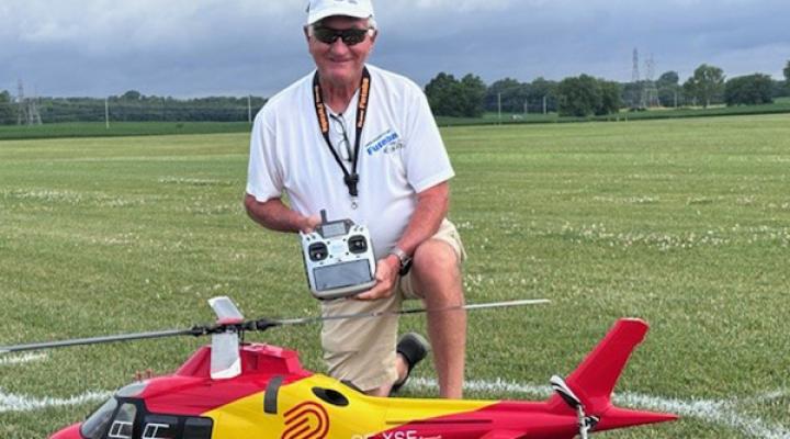 Man kneels on a field, holding a remote control next to a red and yellow model helicopter.