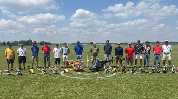 Group of people standing on grass with model helicopters under a blue sky.