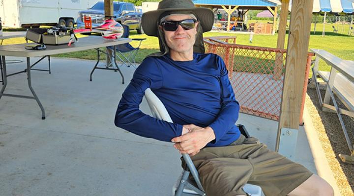 Man in sunglasses and hat, seated outdoors at a picnic area on a sunny day.