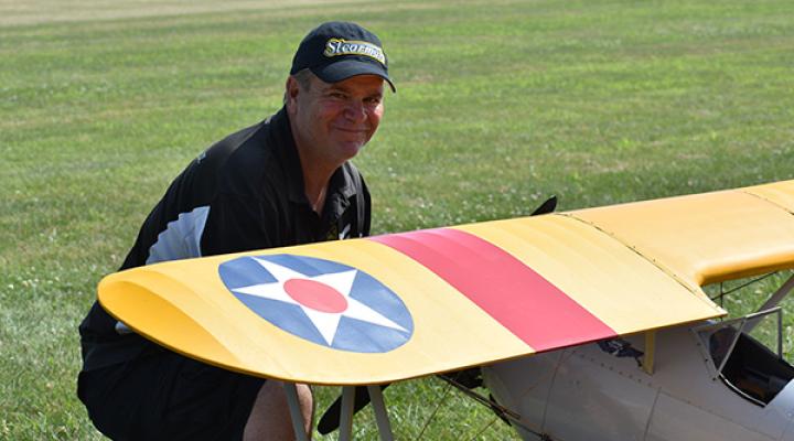 Man smiling next to a model airplane with a yellow wing and emblem on grass.