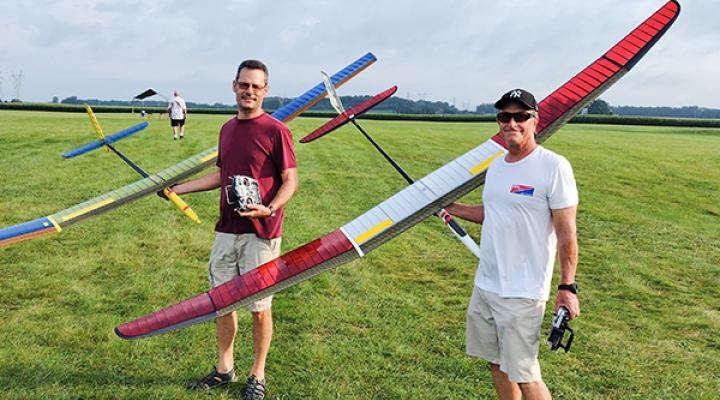 Two men holding colorful model gliders on a grassy field.