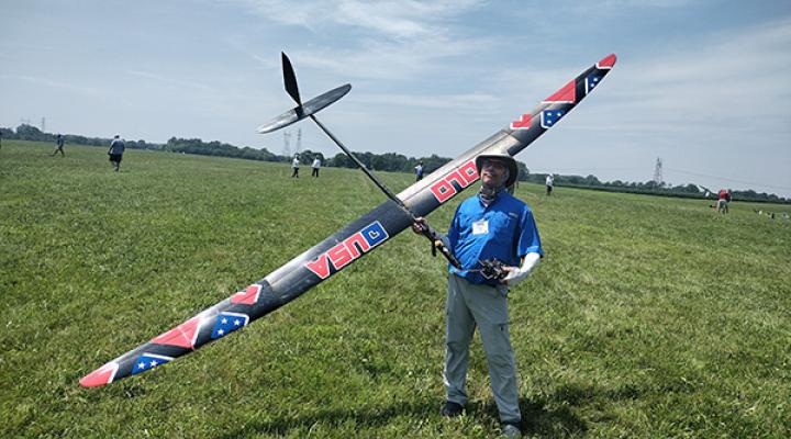 Man holding large model glider on grassy field under blue sky.