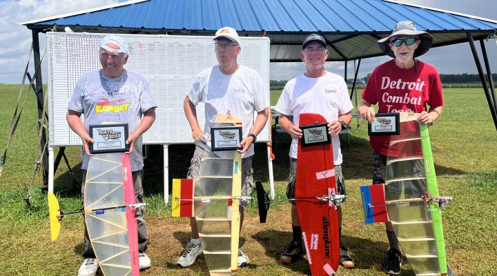 Four people holding model planes and awards, standing on grass near a pavilion.