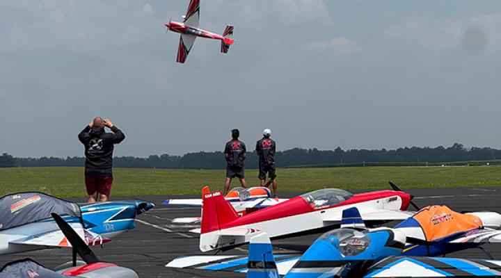 Aerobatic planes on a tarmac, one in flight, with spectators watching.