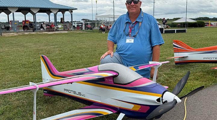 Man posing with colorful model airplane on grassy field.
